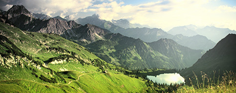 Eine Berglandschaft mit grünen Wiesen und einem See im Tal ist abgebildet.