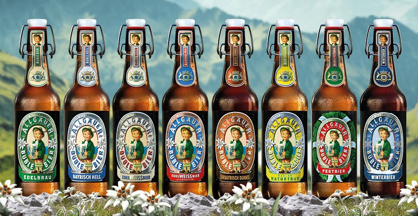 Acht Bierflaschen stehen vor einer Alpenlandschaft, links ein Junge in Tracht hält ein volles Bierkrug.