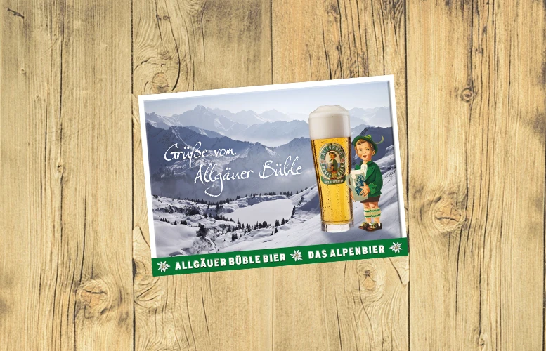 Eine Postkarte mit Bergpanorama und einem BIerglas neben dem eine Figur steht, liegt auf einem Holzuntergrund.
