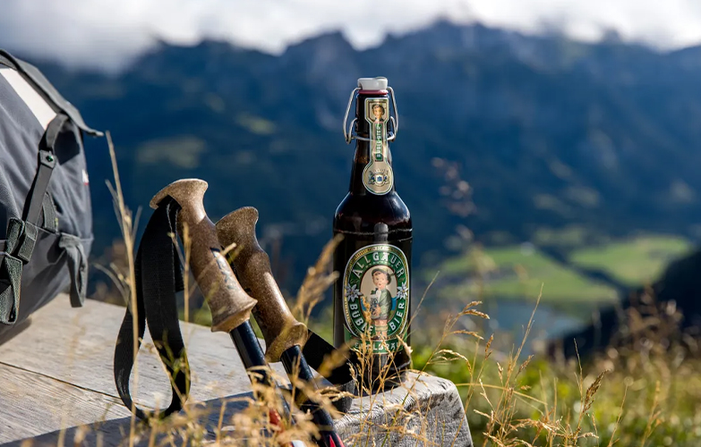 Eine Büble Flasche steht auf eine Holzbank, daneben ein Rucksack und Wanderstöcke,dahinter Bergpanorama.