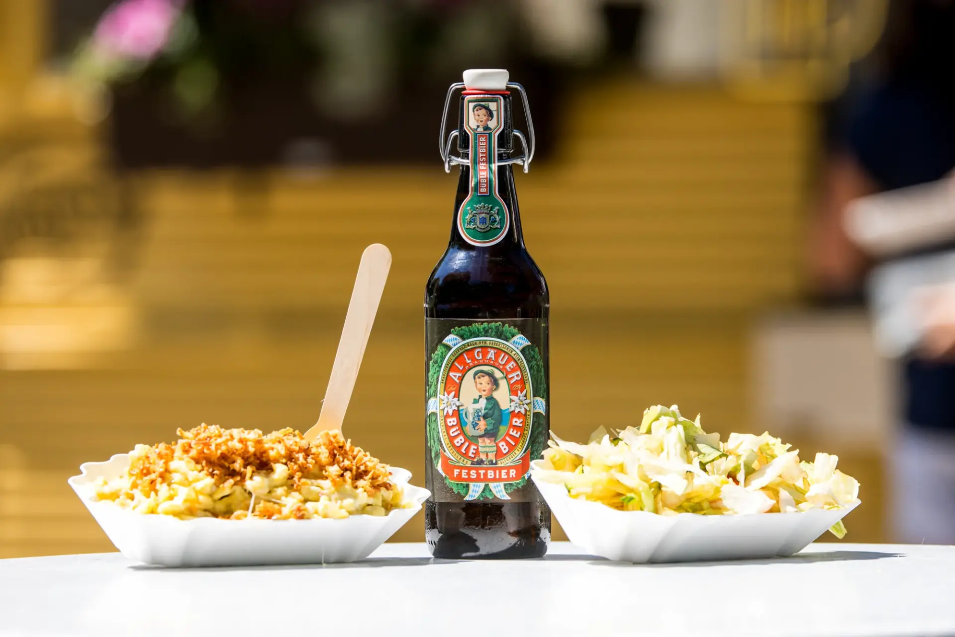 Flasche Allgäuer Büble Festbier steht zwischen Käsespätzle mit Röstzwiebeln und einem Teller Salat.